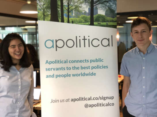 Two students smiling at camera on both sides of a poster with the title  "Apolitical".