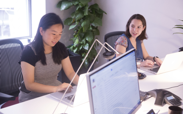 Two women talk while doing work at computers.